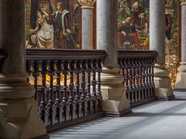 A corridor with stone columns and wooden railings with paintings on the walls at The Mabel Suite Stone Cross Mansion in Ulverston