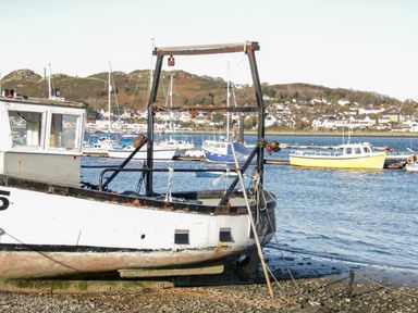 A fishing boat docked on a rocky shore with other boats on the water and a town in the background at The Old Stable in Henryd near Conwy