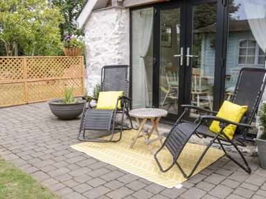 A patio with two black recliner chairs with yellow pillows a small round table and potted plants outside a building with black framed glass doors at The Old Stable in Henryd near Conwy