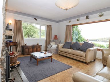 A living room with beige sofas a wooden chest a coffee table and large windows showing greenery and water at The Headland in Llaneilian near Amlwch