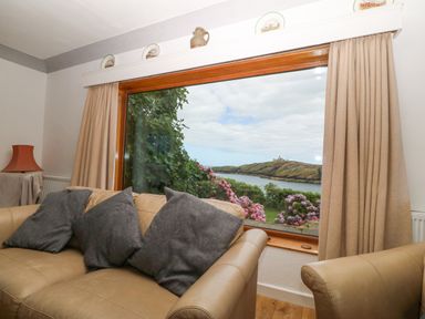 A living room with a beige sofa and gray cushions facing a large window overlooking a garden and water at The Headland in Llaneilian near Amlwch