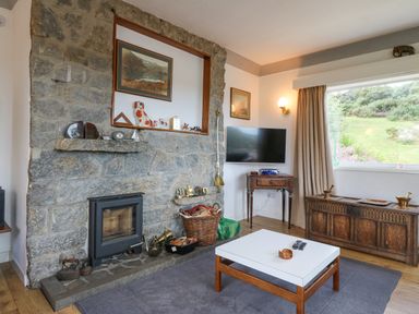 A living room with a stone fireplace wooden coffee table and a large window showing a garden at The Headland in Llaneilian near Amlwch