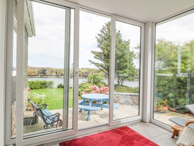 A room with large glass sliding doors opening to a patio with a blue picnic table and benches overlooking a garden and water at The Headland in Llaneilian near Amlwch