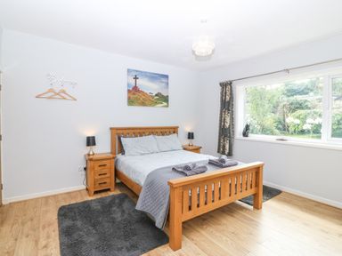 A bedroom with a wooden bed and nightstands with lamps a window showing a garden and rugs on the floor at The Headland in Llaneilian near Amlwch