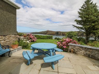 A patio with a round blue wooden picnic table and bench seats overlooking the sea with flowers and trees at The Headland in Llaneilian near Amlwch
