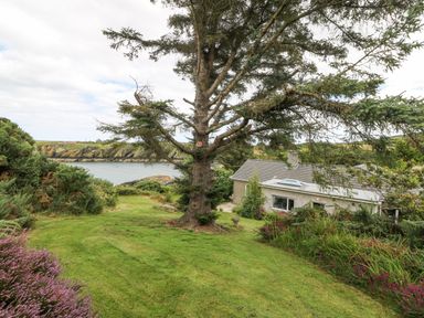 A house with a sloped roof surrounded by trees and shrubs near a body of water at The Headland in Llaneilian near Amlwch