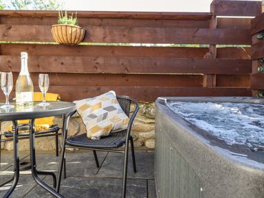 An outdoor patio with a bubbling hot tub a black metal table with two glasses and a bottle and two black chairs one with a patterned cushion at Glyndwr in Betws-Y-Coed