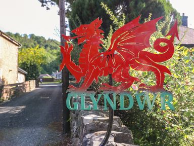 A red dragon metal sign with the word glyndwr on a stone wall by a gravel road with houses and trees in the background at Glyndwr in Betws-Y-Coed