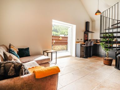 A living room with a sofa next to an open door and a kitchen area with bar stools and a spiral staircase at Glyndwr in Betws-Y-Coed