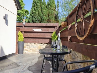An outdoor patio with a round glass table two chairs a bottle of wine two wine glasses wooden fencing and a wagon wheel at Glyndwr in Betws-Y-Coed