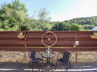 A patio with two black chairs a glass table with a bottle and two glasses a wooden fence and trees in the background at Glyndwr in Betws-Y-Coed