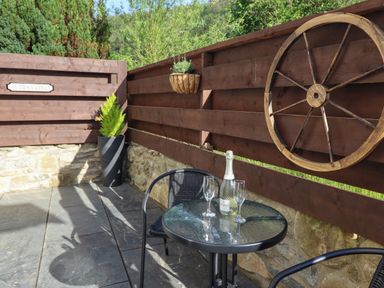 An outdoor patio area with a small round glass table two black chairs a bottle and two glasses on the table wooden fence with a wagon wheel decoration and a plant pot against the wall at Glyndwr in Betws Y Coed