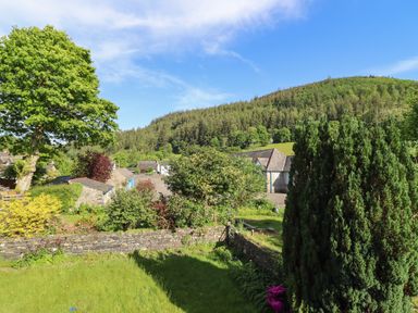 A garden with trees bushes stone walls and houses with a forested hill in the background at Glyndwr in Betws-Y-Coed