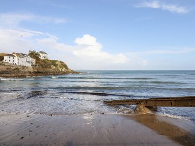 A beach with wet sand and waves near a rocky coastline with houses on a cliff and a rusted metal structure extending into the water at EdyVean Portmellon near Mevagissey