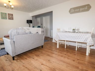 A living room with a gray sofa facing away and a dining table with chairs set for four under a sign on the wall saying Be our guest at EdyVean Portmellon near Mevagissey
