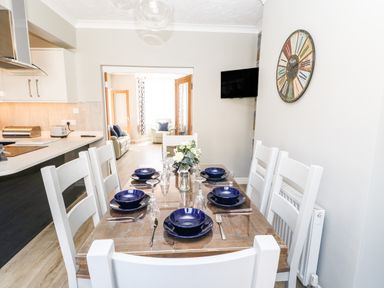A dining area with a wooden table and six white chairs set with blue plates and glassware next to a kitchen with white cabinets and a living room in the background at Lobster Pot Retreat in Newbiggin-By-The-Sea