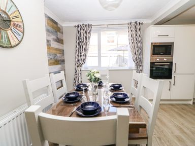 A dining area with a wooden table set with blue dishes and glasses six white chairs a window with curtains and an oven in the corner at Lobster Pot Retreat in Newbiggin-By-The-Sea
