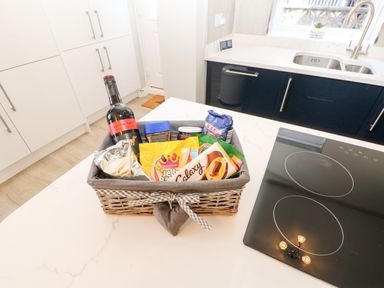 A kitchen countertop with a basket containing a bottle of wine and assorted snacks near a stove and sink at Lobster Pot Retreat in Newbiggin-By-The-Sea
