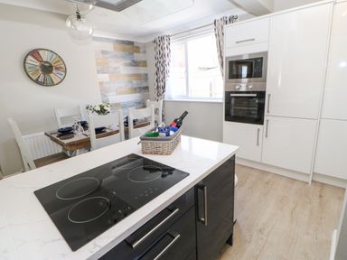 A kitchen with a stove on an island a dining table with chairs and a wall clock at Lobster Pot Retreat in Newbiggin-By-The-Sea