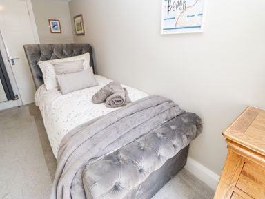 A bedroom with a single bed with gray tufted headboard and footboard a wooden nightstand and framed pictures on the wall at Lobster Pot Retreat in Newbiggin-By-The-Sea