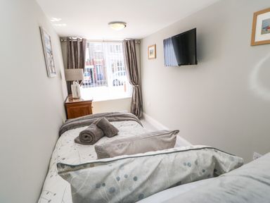 A bedroom with a single bed a wooden nightstand lamp wall mounted tv and window with curtains at Lobster Pot Retreat in Newbiggin-By-The-Sea