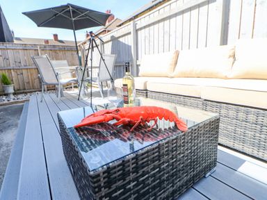 An outdoor patio with a wicker sofa a glass table with a lobster a bottle of wine and a glass a dining table with chairs and an umbrella at Lobster Pot Retreat in Newbiggin-By-The-Sea