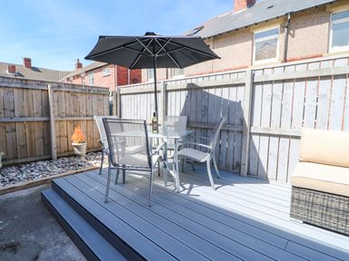 An outdoor patio area with a glass table four chairs and a black umbrella next to a wicker bench with beige cushions at Lobster Pot Retreat in Newbiggin-By-The-Sea