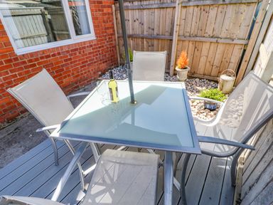 An outdoor patio area with a glass table and four chairs a bottle and glass on the table against a brick wall and wooden fence at Lobster Pot Retreat in Newbiggin-By-The-Sea