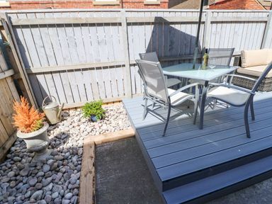 An outdoor patio with a glass table and four chairs on a wooden deck next to a gravel area with plants and a watering can