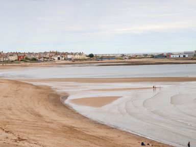 A sandy beach with shallow water and people walking near the shore with a row of houses in the background at Lobster Pot Retreat in Newbiggin-By-The-Sea