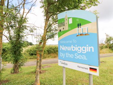 A welcome sign for Newbiggin by the Sea near trees and a road