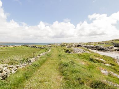 A grassy pathway with stone walls leading to a coastal area with rocks and the ocean at Ti Tom Narta in Trabane near Lettermore County Galway