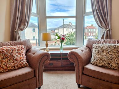 Two armchairs with cushions facing a window with curtains and a table with a lamp and flowers at Grove House in Egremont