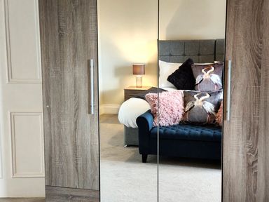 A bedroom with a bed a side table with a lamp and a couch reflected in a wardrobe mirror at Grove House in Egremont