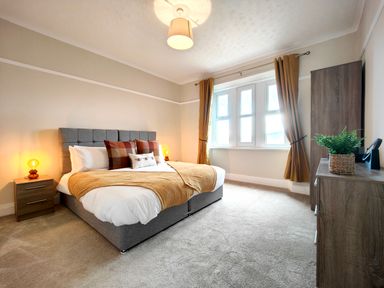 A bedroom with a gray bed with pillows and a blanket beside nightstands with lamps and a window with curtains at Grove House in Egremont