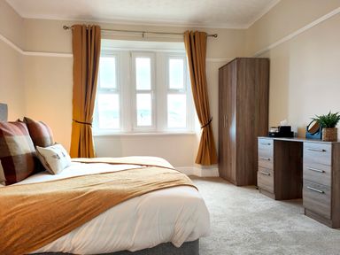 A bedroom with a bed, brown curtains, wardrobe, desk with drawers, and a plant at Grove House in Egremont