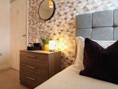 A bedroom corner with a bed pillow bedside table drawers lamp plant and round mirror on patterned wall at Grove House in Egremont