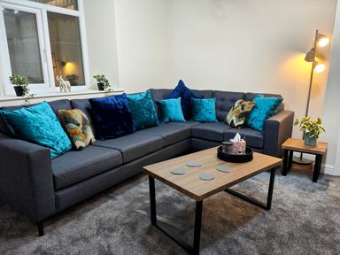 A living room with a gray sectional sofa with teal and patterned cushions a wooden coffee table with coasters and a side table with a plant at Grove House in Egremont
