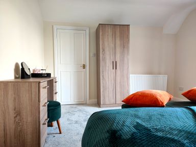 A bedroom with a wooden wardrobe a wooden dresser a round green stool an orange pillow and a blue blanket at Grove House in Egremont