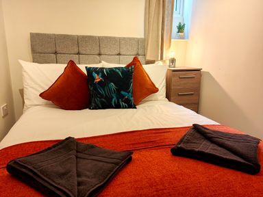 A bed with white sheets, orange and patterned pillows, orange blanket, black towels and a wooden nightstand with a lamp and a plant at Grove House in Egremont