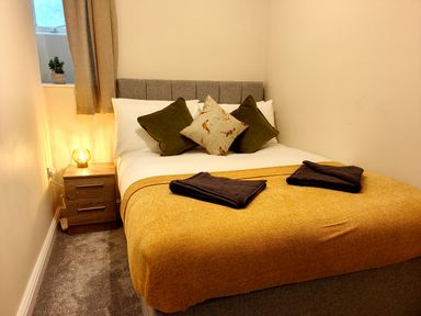 A bedroom with a bed covered with a yellow throw and pillows beside a wooden nightstand with a lamp at Grove House in Egremont