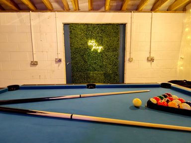 A pool table with pool cues and balls in a room with a green wall feature and a neon sign that says lets play at Grove House in Egremont