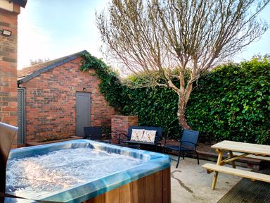 An outdoor patio with a hot tub seating area with chairs and a bench and a wooden picnic table near a brick shed and a leafless tree with greenery in the background at Grove House in Egremont