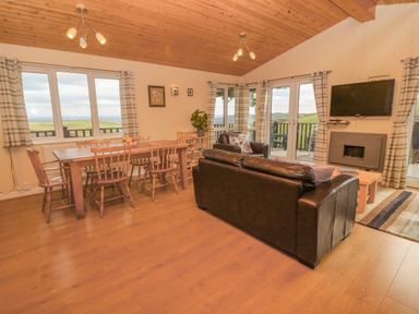 A living room with a wooden dining table and chairs a black leather sofa a mounted TV and windows showing a countryside view at Lodge 23 in Torpoint