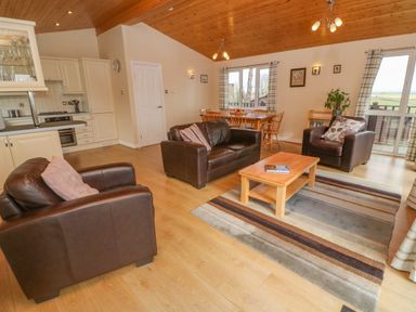 An open plan living room and kitchen with brown leather sofas a wooden coffee table a dining table and windows with curtains at Lodge 23 in Torpoint
