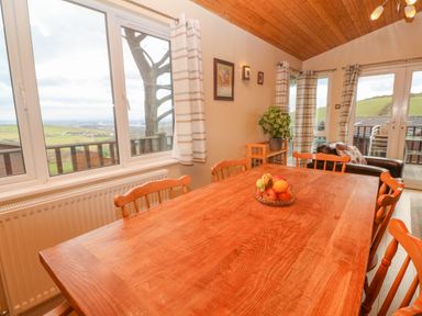A dining area with a wooden table and chairs a fruit bowl on the table plants and framed pictures on the wall windows with a view of green hills and a deck outside at Lodge 23 in Torpoint