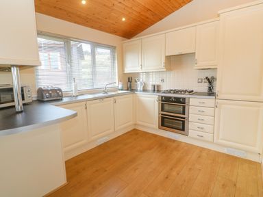 A kitchen with cream cabinets, wood flooring, a gas stove with oven, a microwave, toaster, sink under a window with blinds and a wooden ceiling at Lodge 23 in Torpoint