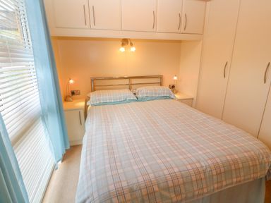 A bedroom with a double bed with plaid bedding two bedside tables with lamps overhead cabinets and a window with blue curtains at Lodge 23 in Torpoint