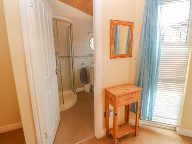 A small wooden table with a drawer and a wall mirror beside a window with blue curtains next to a bathroom door with a shower at Lodge 23 in Torpoint