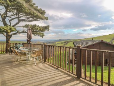 A wooden deck with a glass table and chairs and a closed umbrella overlooking rolling green hills and a wooden cabin at Lodge 23 in Torpoint